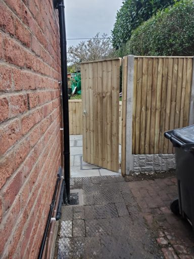 Narrow pathway between a brick wall and wooden fence, leading to a garden gate.