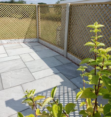 Patio area with stone flooring and a trellis fence, surrounded by greenery.