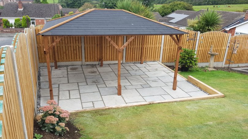 Wooden gazebo with a slate roof on a stone-paved patio in a fenced garden area.