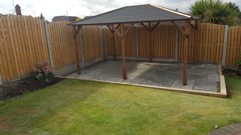 A wooden garden gazebo on a concrete base, surrounded by grass and a wooden fence.