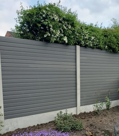 Grey vertical fence with a lush green hedge and flowers on top.