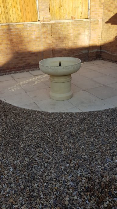 A stone fountain in a courtyard with a gravel surface and wooden backdrop.