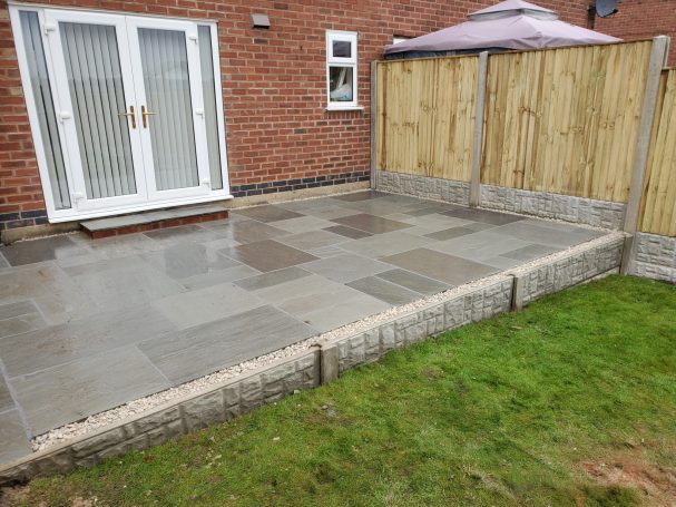 Paved outdoor area with stone tiles, bordered by a wooden fence and lawn.