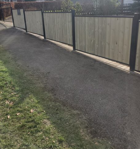 Pathway beside a wooden fence, with grass on one side and a concrete surface.