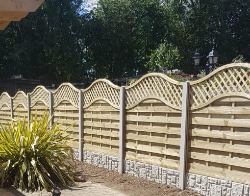 A decorative wooden fence with a wavy top and concrete posts, surrounded by greenery.