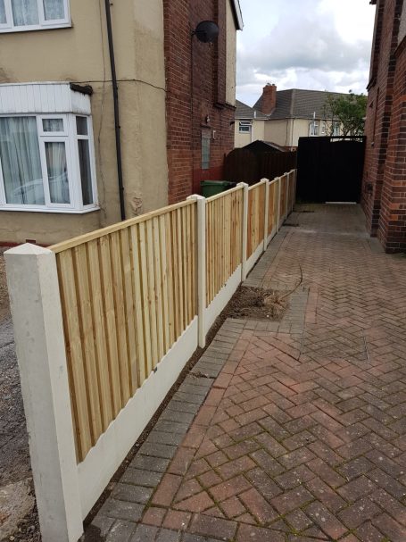 Narrow paved pathway with a wooden fence on one side and a darkened area at the end.