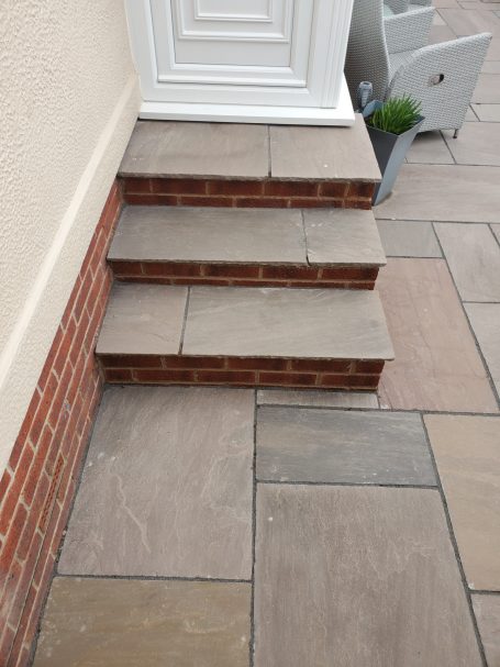 Three stone steps leading up to a white door, with surrounding patterned paving.