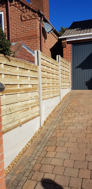 A wooden fence along a paved driveway with a house and garage in the background.