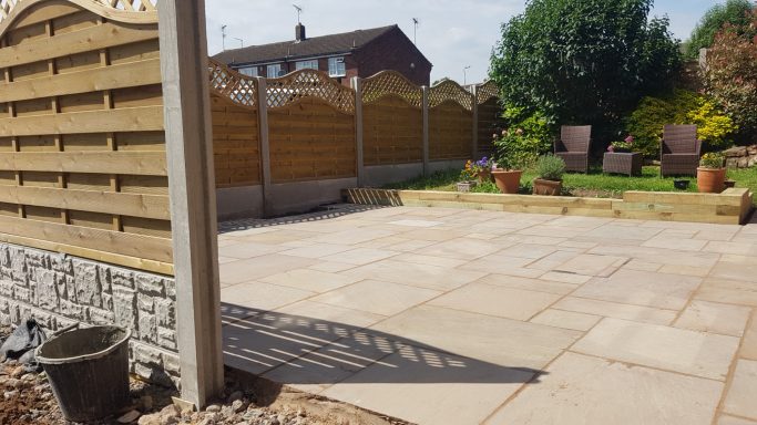 Patio area with paved stones, garden furniture, and planted pots surrounded by wooden fences.