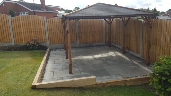 Wooden gazebo on a patio with a grass lawn and wooden fence in the background.
