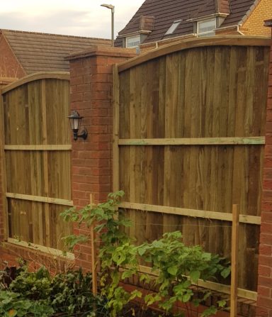 Wooden fence with arched tops, separating garden from a brick wall, with plants in front.