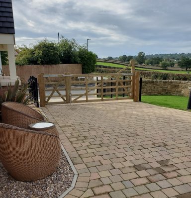 A gravel driveway with a wooden gate and two seating areas, surrounded by greenery.