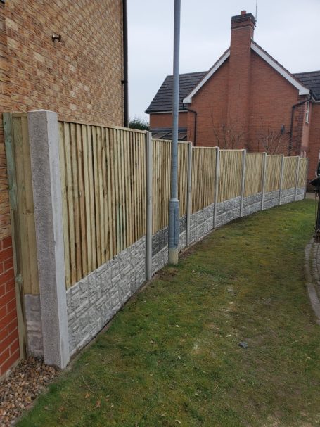 Curved wooden and concrete fence along a grassy pathway beside residential buildings.
