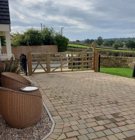 A wooden gate opens to a scenic landscape; stone path and chairs are in the foreground.