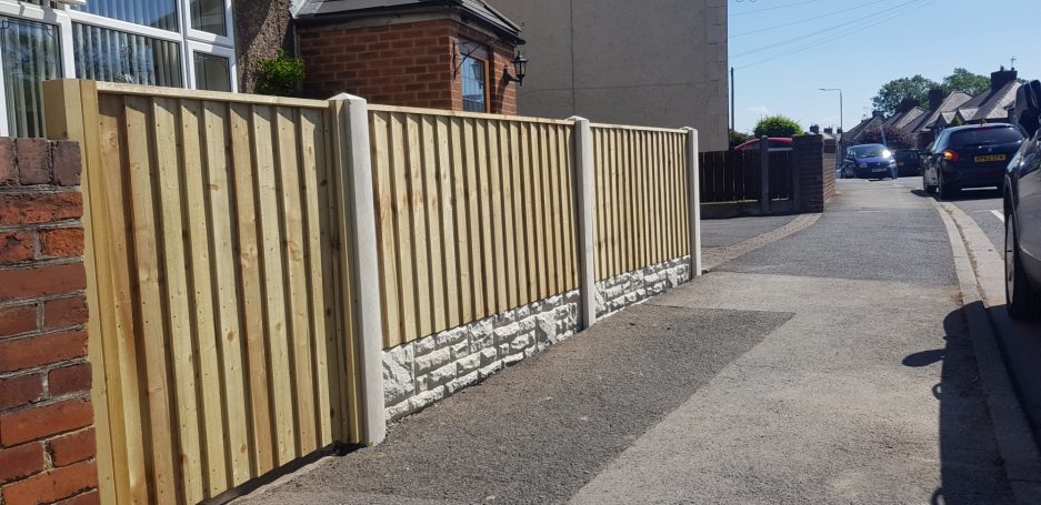 Wooden fence alongside a road with parked cars and buildings in the background.