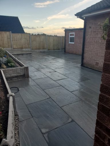 Paved patio area beside a house, with a wooden fence and garden bed.