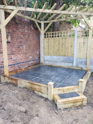 Patio area with wooden framing and brick wall, surrounded by earth and fencing.