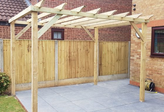Wooden pergola frame in a garden with a paved area and wooden fence backdrop.