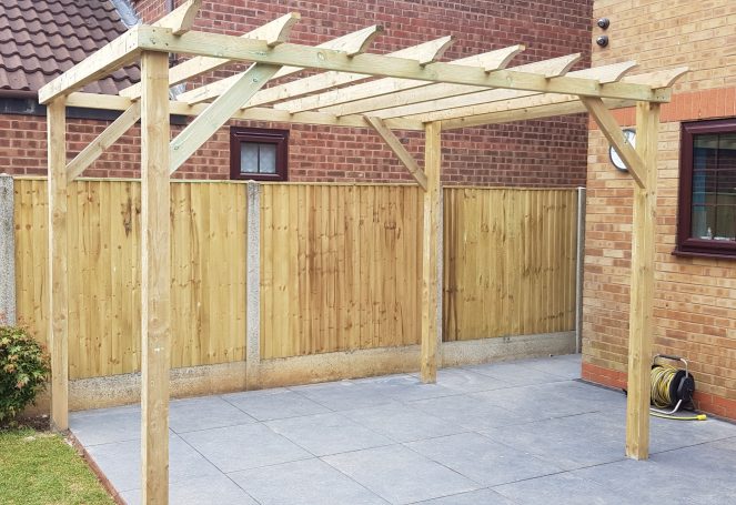 Wooden pergola structure in a garden with a paved area and wooden fencing in the background.