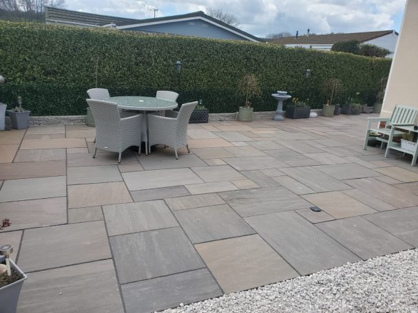 Paved outdoor patio with a table, chairs, and green hedges in the background.